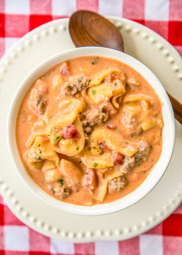 bowl of tomato soup with tortellini and italian sausage on a plate with a spoon on top of a red checked tablecloth