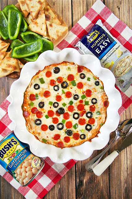 Baked Pizza Hummus Dip - quick homemade pizza hummus topped with mozzarella, parmesan and your favorite pizza toppings. Start with garbanzo beans and a pouch of Bush's Classic Hummus Made Easy, add tomato paste, garlic, Italian Seasoning and parmesan cheese. Bake for 15 minutes. Serve with bell pepper slices and pita chips. Great for a quick party appetizer or alternative for pizza night!