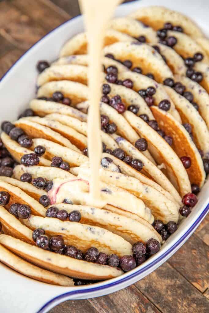 pouring egg custard over blueberries and pancakes in a baking dish