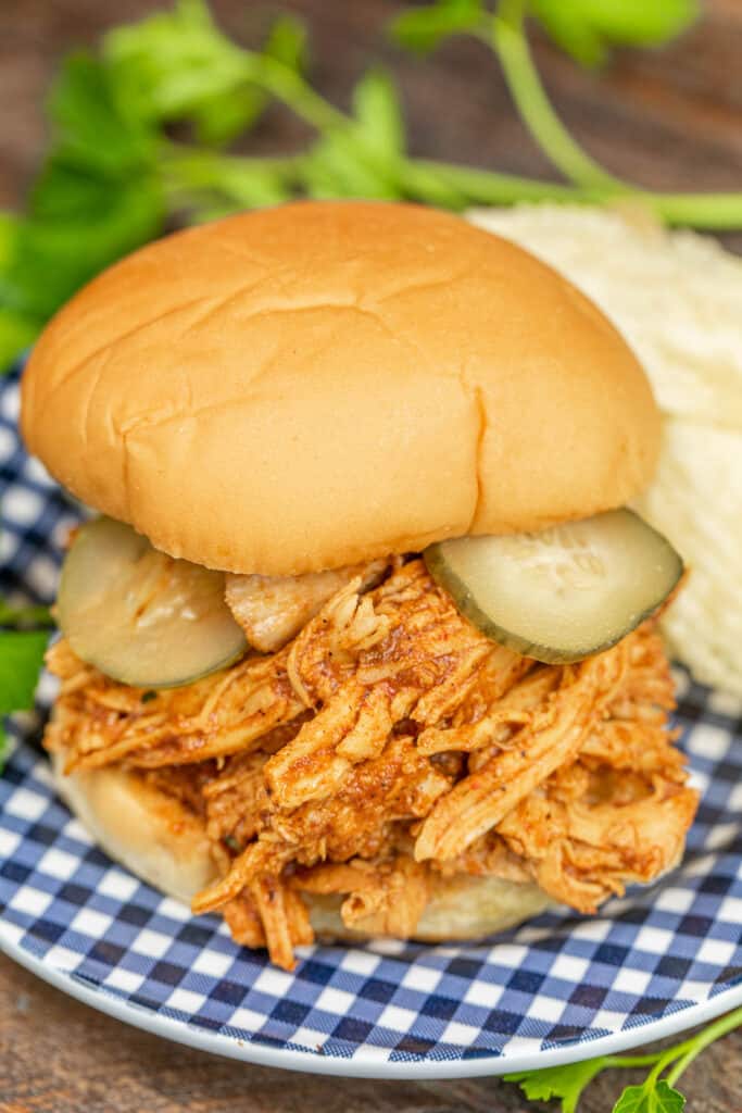 shredded bbq chicken sandwich topped with pickles on a blue checkered late with chips