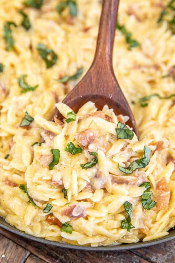 scooping creamy orzo with prosciutto and basil from skillet