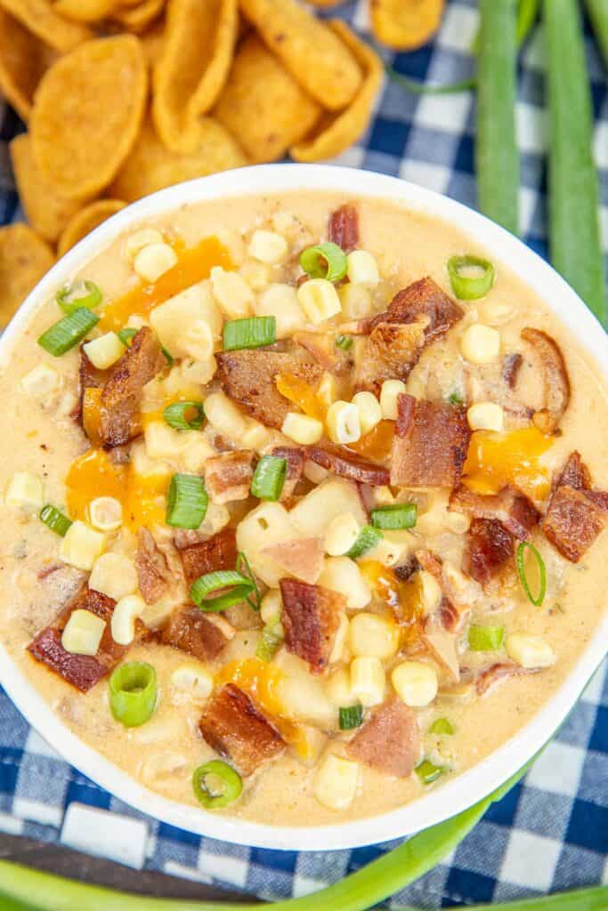 bowl of corn chowder with fritos and green onions sitting on a blue and white checked tablecloth