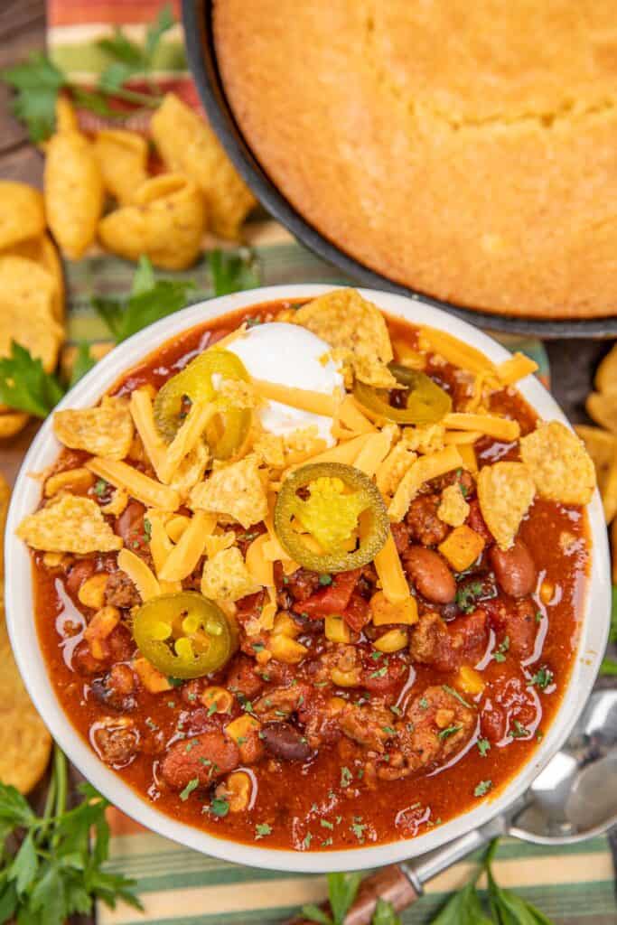 bowl of chili topped with sour cream, cheese, jalapenos, and crusted fritos in front of a pan of cornbread