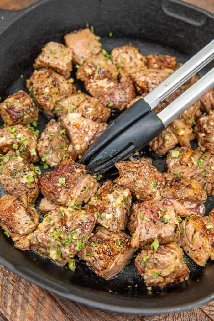cooking steak bites in a skillet with tongs